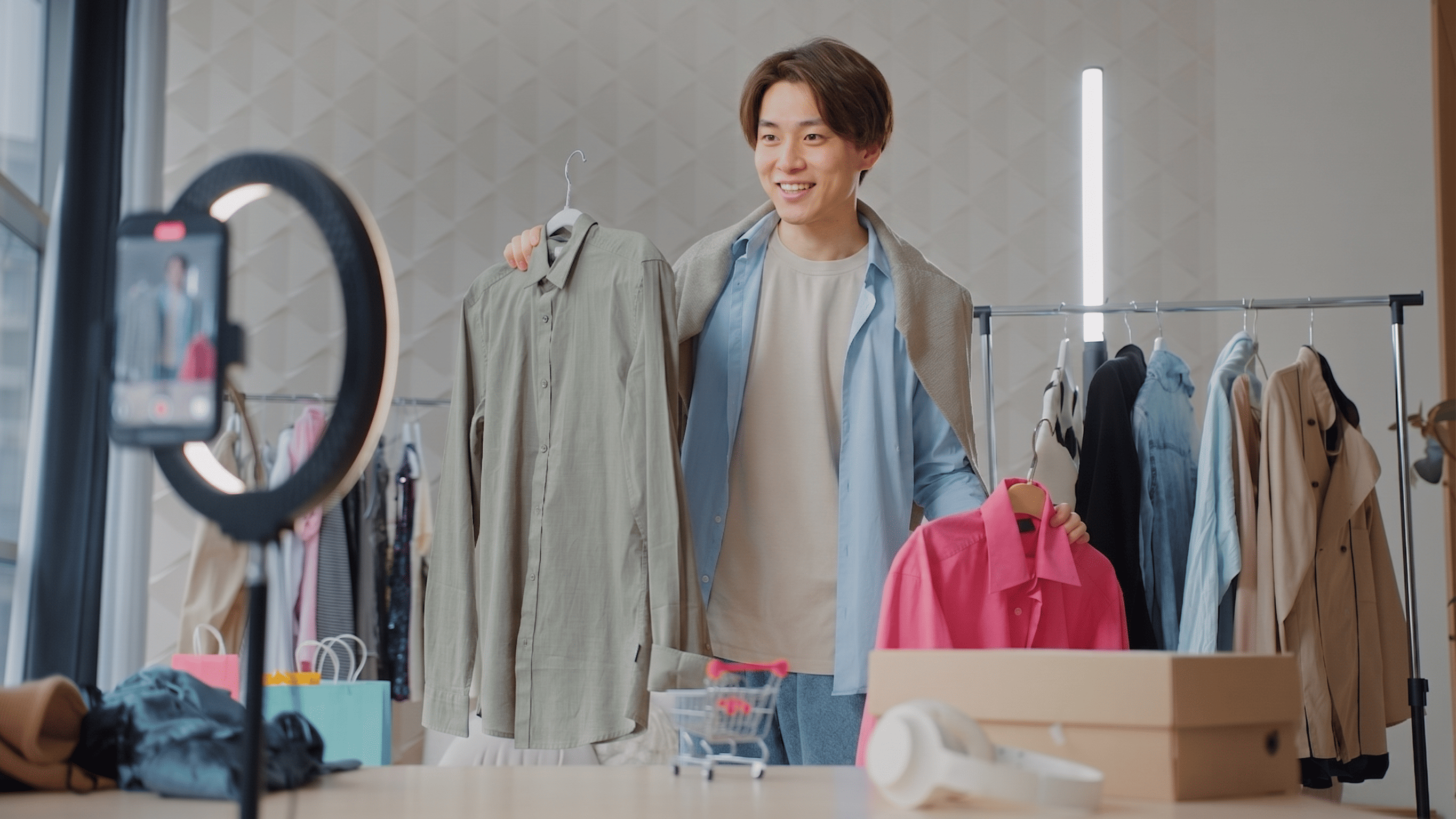 たくさんの洋服が並んでいる部屋で男性がスマートフォンを使ってカーキのシャツと赤色のシャツを紹介しています